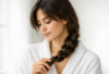 young woman in her late 20s with long dark brown hair in a soft loose braid, wearing a white cotton robe, gently touching the end of her braid and looking down with a calm, slightly thoughtful expression, natural morning light